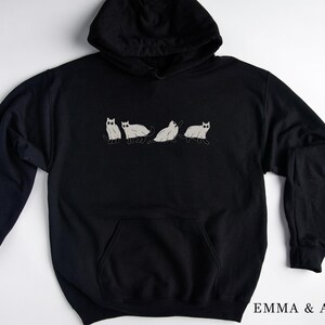 Puede incluir: Sudadera con capucha negra con un gr&aacute;fico de cinco gatos blancos en fila. El texto "EMMA & ANNA" est&aacute; impreso en la esquina inferior derecha de la sudadera.