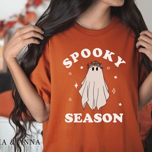 May include: A burnt orange t-shirt with a white graphic of a ghost wearing a flower crown and the text "Spooky Season".