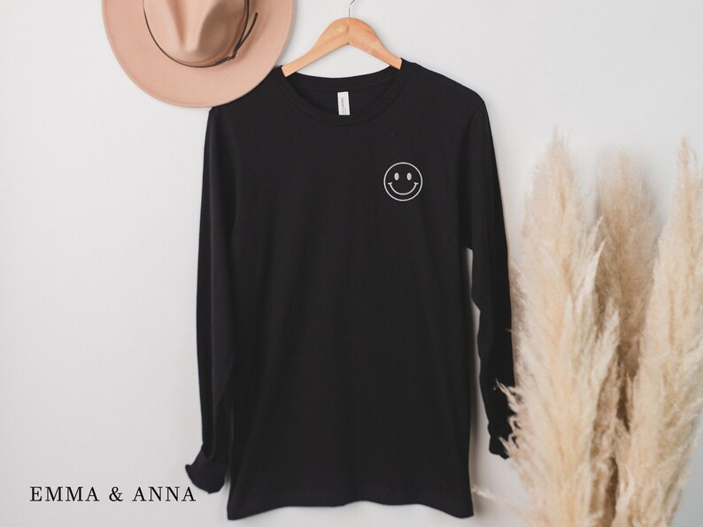 Puede incluir: Camiseta negra de manga larga con un gr&aacute;fico de carita sonriente blanca en el pecho izquierdo. La camiseta est&aacute; colgada de una percha de madera. Un sombrero color canela es visible en el fondo. El texto "EMMA & ANNA" est&aacute; impreso abajo a la izquierda.