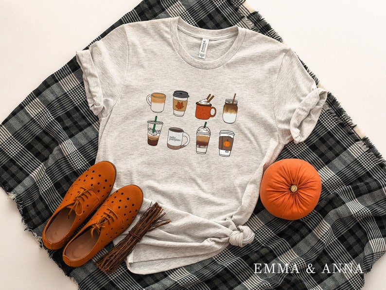 Może przedstawiać: Szary T-shirt z grafiką przedstawiającą r&oacute;żne napoje kawowe, w tym latte macchiato, cappuccino i kawę mrożoną. Na dole koszulki nadrukowany jest tekst "EMMA & ANNA".