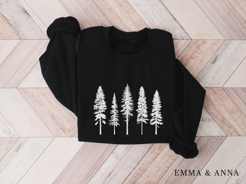 Puede incluir: Sudadera negra con un gr&aacute;fico blanco de cinco pinos. El texto "EMMA & ANNA" est&aacute; impreso en la esquina inferior derecha de la sudadera.