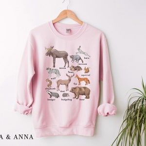 May include: Pink sweatshirt with a graphic print of various woodland animals, including a moose, deer, fox, squirrel, raccoon, badger, hedgehog, and bear. The animals are labeled with their names.