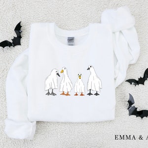 May include: White sweatshirt with a graphic of four ghost ducks. The ducks are wearing white sheets and have orange beaks. The text "EMMA & ANNA" is printed on the bottom of the sweatshirt.