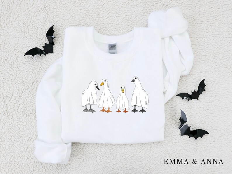 May include: White sweatshirt with a graphic of four ghost ducks. The ducks are wearing white sheets and have orange beaks. The text "EMMA & ANNA" is printed on the bottom of the sweatshirt.
