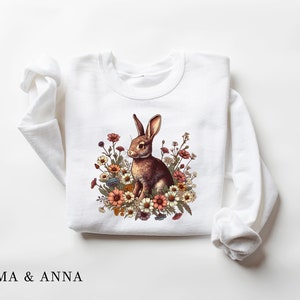 Pu&ograve; includere: Felpa bianca con un coniglio marrone circondato da fiori colorati. Il disegno &egrave; centrato sulla parte anteriore del top a maniche lunghe. Il testo "EMMA & ANNA" &egrave; stampato in basso.