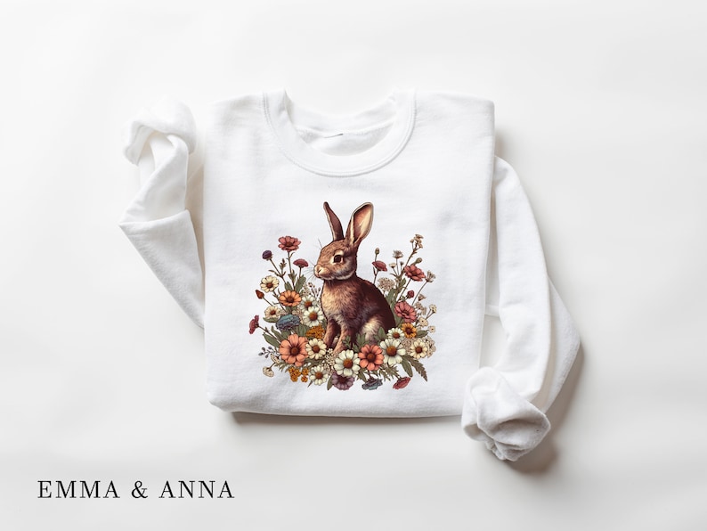 Pu&ograve; includere: Felpa bianca con un coniglio marrone circondato da fiori colorati. Il disegno &egrave; centrato sulla parte anteriore del top a maniche lunghe. Il testo "EMMA & ANNA" &egrave; stampato in basso.