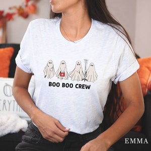 Puede incluir: Una camiseta blanca con un gr&aacute;fico de cinco fantasmas vestidos como profesionales m&eacute;dicos. El texto "BOO BOO CREW" est&aacute; impreso debajo de los fantasmas.