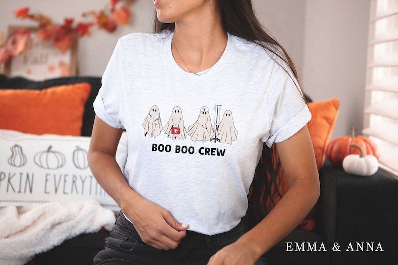 Puede incluir: Una camiseta blanca con un gr&aacute;fico de cinco fantasmas vestidos como profesionales m&eacute;dicos. El texto "BOO BOO CREW" est&aacute; impreso debajo de los fantasmas.