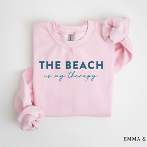 以下が含まれることがあります： 濃い青色の文字で「THE BEACH is my therapy」と書かれたピンクのクルーネックのトレーナー