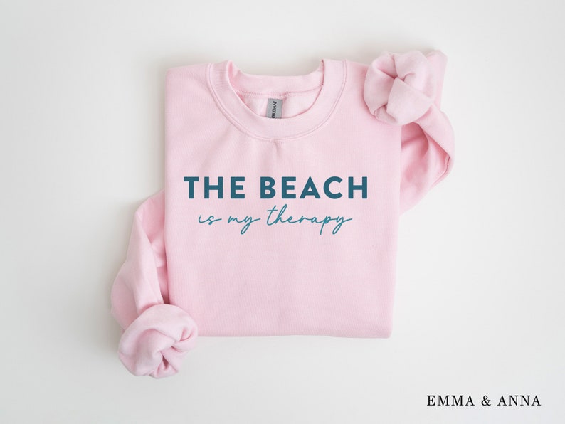 以下が含まれることがあります： 濃い青色の文字で「THE BEACH is my therapy」と書かれたピンクのクルーネックのトレーナー