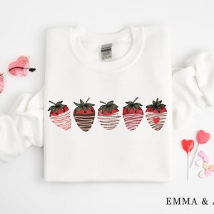 Puede incluir: Sudadera blanca con un gr&aacute;fico de cinco fresas cubiertas de chocolate. Las fresas est&aacute;n dispuestas en una fila y tienen rayas blancas y marrones.