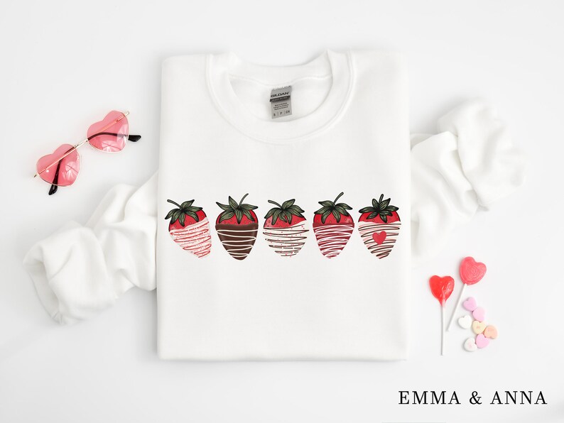 Puede incluir: Sudadera blanca con un gr&aacute;fico de cinco fresas cubiertas de chocolate. Las fresas est&aacute;n dispuestas en una fila y tienen rayas blancas y marrones.