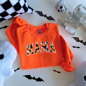 Op de afbeelding: Fel oranje sweatshirt met het woord "MAMA" in een lettertype in collegestijl, met een bloemen- en pompoenpatroon. Halloween-decoraties zijn onder meer vleermuizen, spoken en een skelet.