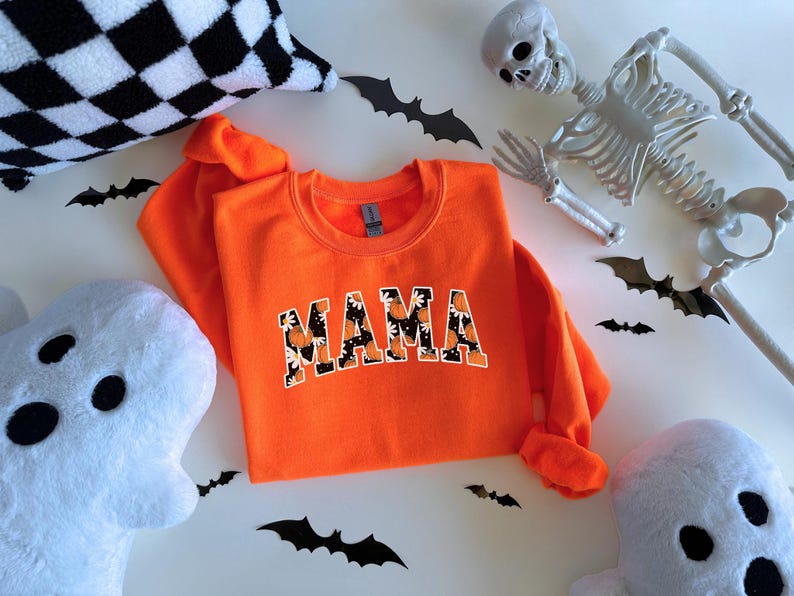 Op de afbeelding: Fel oranje sweatshirt met het woord "MAMA" in een lettertype in collegestijl, met een bloemen- en pompoenpatroon. Halloween-decoraties zijn onder meer vleermuizen, spoken en een skelet.