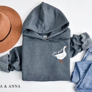 K&ouml;nnte beinhalten: Ein dunkelgrauer Kapuzenpullover mit dem Text "silly goose" und einer wei&szlig;en G&auml;nseillustration auf der Vorderseite.