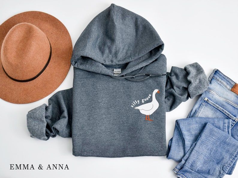 K&ouml;nnte beinhalten: Ein dunkelgrauer Kapuzenpullover mit dem Text "silly goose" und einer wei&szlig;en G&auml;nseillustration auf der Vorderseite.