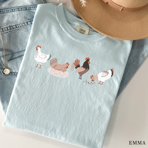 May include: A light blue t-shirt with a graphic of four chickens. The chickens are white, brown, and black with red combs. The text "COMFORT  COLOR" is on the shirt.