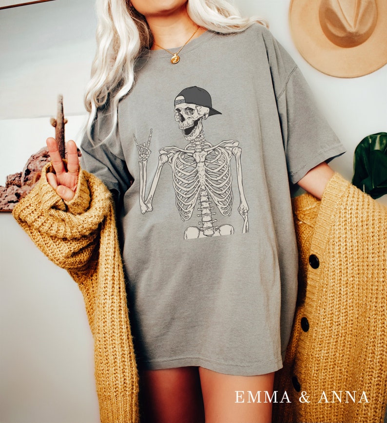 May include: A gray oversized t-shirt featuring a skeleton wearing a baseball cap and giving a peace sign. The skeleton is in a relaxed pose and has a friendly expression. The shirt is made of a soft, comfortable material and has a relaxed fit.
