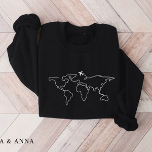 May include: Black sweatshirt with a white outline of a world map and two airplanes. The sweatshirt is folded and laid flat on a wooden surface.