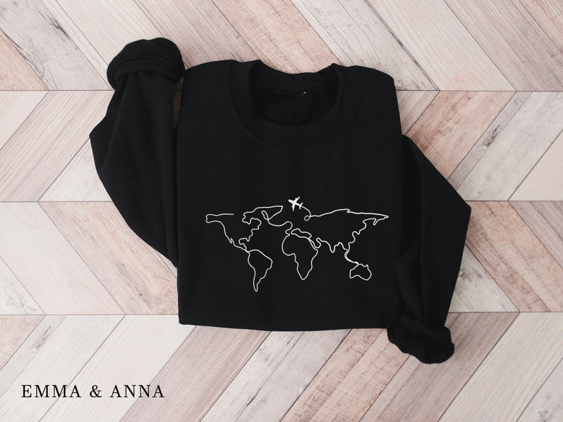 May include: Black sweatshirt with a white outline of a world map and two airplanes. The sweatshirt is folded and laid flat on a wooden surface.