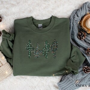 May include: A green sweatshirt with a design of four cactus plants with white and pink flowers. The sweatshirt is on a white fuzzy surface with pine cones and string lights.