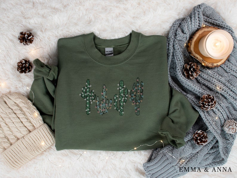 May include: A green sweatshirt with a design of four cactus plants with white and pink flowers. The sweatshirt is on a white fuzzy surface with pine cones and string lights.