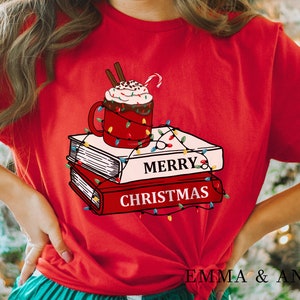 May include: Red t-shirt with a graphic of a mug of hot chocolate with whipped cream and a candy cane on top of a stack of books with Christmas lights wrapped around them. The text "Merry Christmas" is printed on the books.