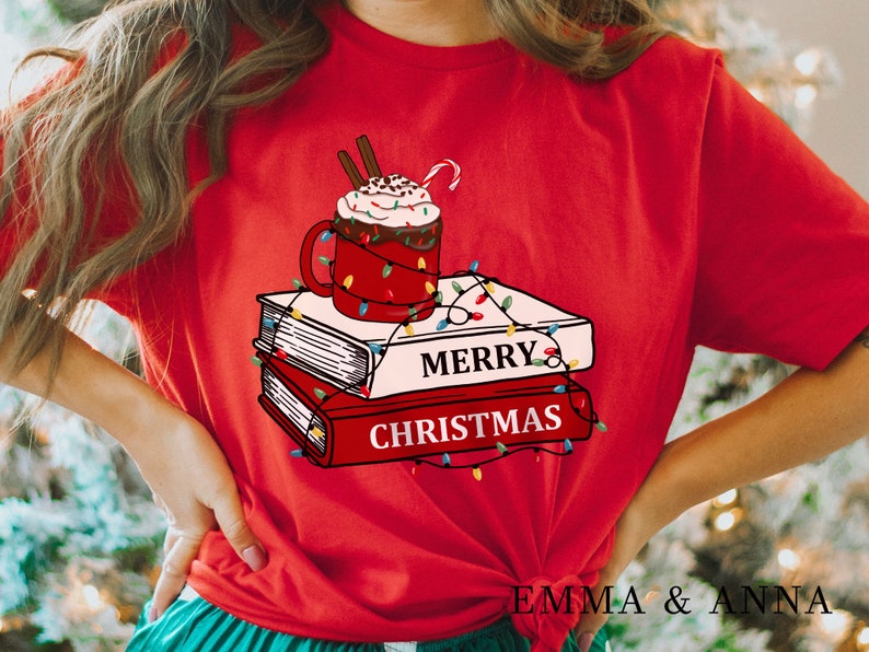 May include: Red t-shirt with a graphic of a mug of hot chocolate with whipped cream and a candy cane on top of a stack of books with Christmas lights wrapped around them. The text "Merry Christmas" is printed on the books.