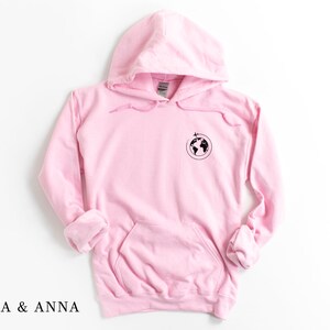 May include: Pink hoodie with a black globe and airplane graphic on the front. The hoodie has a drawstring hood and a kangaroo pocket.