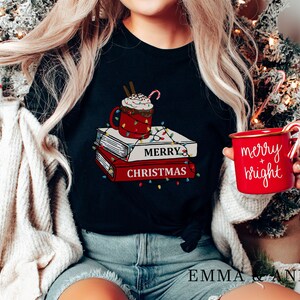 May include: A black t-shirt with a graphic of a mug of hot chocolate with whipped cream and a candy cane on top of a stack of books with the text "Merry Christmas" and lights around the books. The person is holding a red mug with the text "merry + bright" and a candy cane.