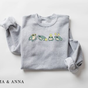 Puede incluir: Una sudadera gris con un gr&aacute;fico de cuatro ranas con sombreros y sosteniendo tr&eacute;boles. El texto "EMMA & ANNA" est&aacute; impreso debajo de la imagen.