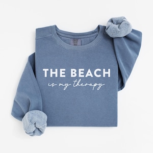 May include: A blue sweatshirt with the phrase "THE BEACH is my therapy" in white lettering. The long sleeves are cuffed, and a matching scrunchie is on the shoulder. The sweatshirt is a solid color and has a casual style.