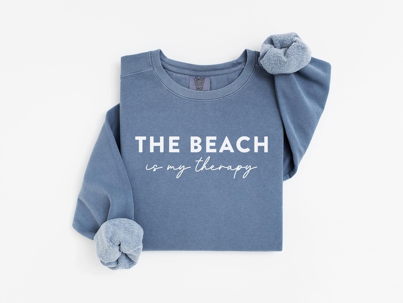 Pu&ograve; includere: Una felpa blu con la scritta bianca "THE BEACH is my therapy". Le maniche sono arrotolate e un elastico per capelli abbinato &egrave; sulla spalla. La felpa &egrave; di colore pieno e ha uno stile casual.