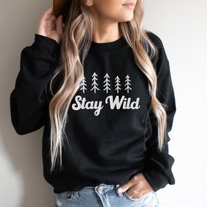 K&ouml;nnte beinhalten: Schwarzer Pullover mit einem wei&szlig;en Aufdruck einer Reihe von Kiefern und dem Text "Stay Wild".