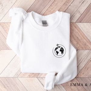 May include: White sweatshirt with a black graphic of a globe with airplanes flying around it. The text "EMMA & ANNA" is printed on the bottom right corner of the image.