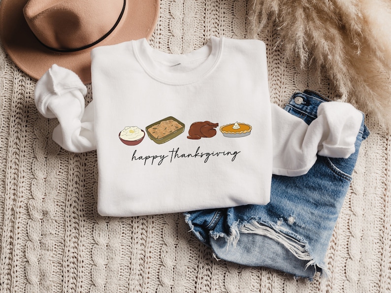 K&ouml;nnte beinhalten: Wei&szlig;es Sweatshirt mit einem Thanksgiving-Motiv mit einem Truthahn, Kartoffelp&uuml;ree, F&uuml;llung und K&uuml;rbiskuchen. Der Text "happy thanksgiving" ist unter den Speisen gedruckt.