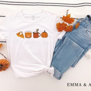 May include: White t-shirt with a graphic of a pumpkin spice latte, pumpkin pie, and a candle. The shirt is folded and laid on a white and black striped surface with a pair of blue jeans and fall leaves.
