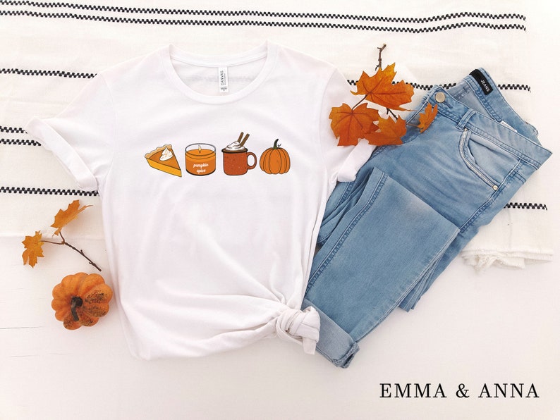 May include: White t-shirt with a graphic of a pumpkin spice latte, pumpkin pie, and a candle. The shirt is folded and laid on a white and black striped surface with a pair of blue jeans and fall leaves.