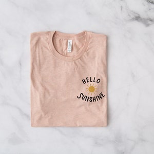 May include: A folded pink t-shirt with a black graphic that says "Hello Sunshine" with a yellow sun graphic.