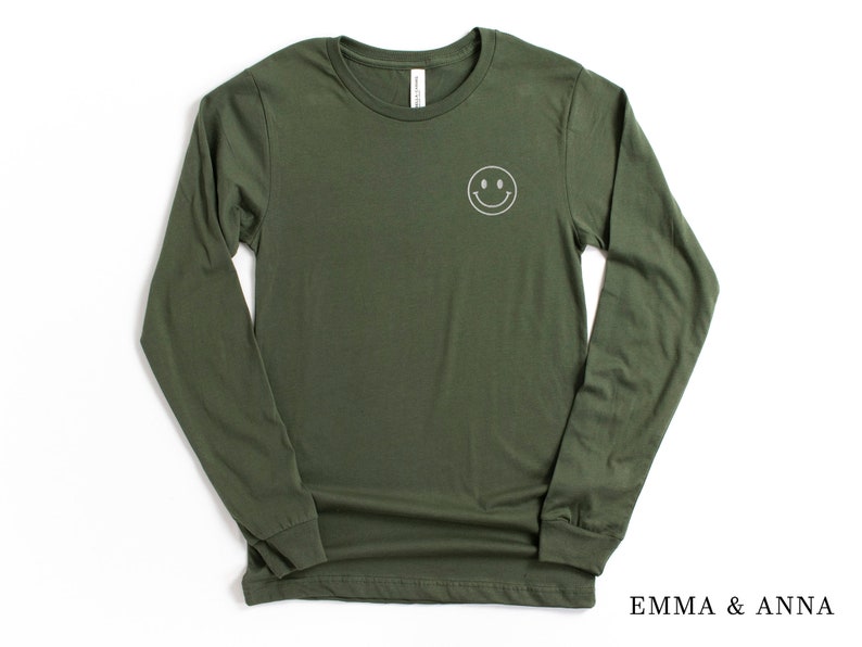 Puede incluir: Camiseta de manga larga verde oliva con una carita sonriente blanca bordada en el pecho. La camiseta tiene cuello redondo y mangas largas. El nombre de la marca "EMMA & ANNA" est&aacute; impreso en negro en la parte inferior derecha.