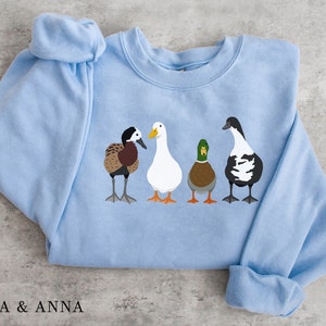 Peut inclure: Un sweat-shirt bleu clair avec un motif de quatre canards align&eacute;s. Les canards sont brun et blanc, blanc, vert et brun, et noir et blanc.