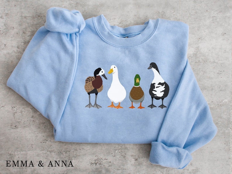 Peut inclure: Un sweat-shirt bleu clair avec un motif de quatre canards align&eacute;s. Les canards sont brun et blanc, blanc, vert et brun, et noir et blanc.