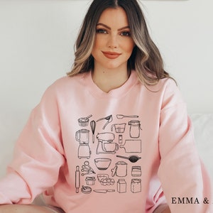 May include: A pink sweatshirt with a black graphic print of various kitchen tools and utensils. The text "EMMA & ANNA" is printed on the bottom right corner of the sweatshirt.