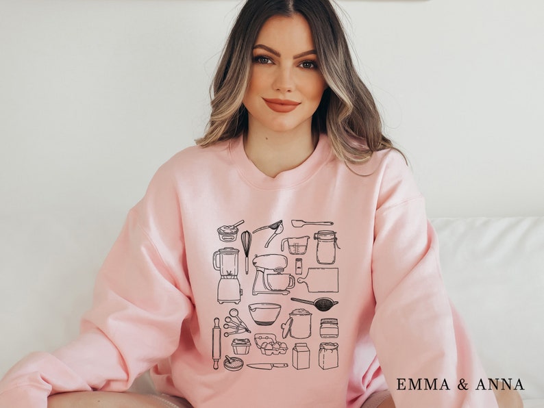 May include: A pink sweatshirt with a black graphic print of various kitchen tools and utensils. The text "EMMA & ANNA" is printed on the bottom right corner of the sweatshirt.