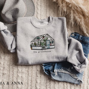 May include: A light grey sweatshirt with a greenhouse illustration and the words "the greenhouse". The image shows a detailed drawing of a greenhouse filled with plants. The sweatshirt is styled with denim shorts and a grey hat.