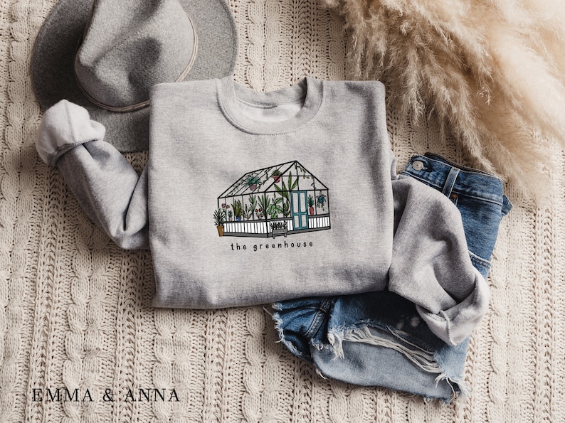 May include: A light grey sweatshirt with a greenhouse illustration and the words "the greenhouse". The image shows a detailed drawing of a greenhouse filled with plants. The sweatshirt is styled with denim shorts and a grey hat.