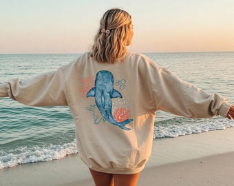May include: Beige sweatshirt with a blue whale design, coral, and bubbles. The whale has white speckles. The sweatshirt is worn on a beach with the sea in the background.