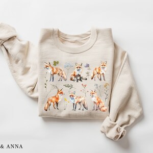 May include: A beige sweatshirt with a print of multiple red foxes and wildflowers. The foxes are in various poses, including sitting, standing, and walking. The wildflowers are in shades of pink, purple, and yellow.