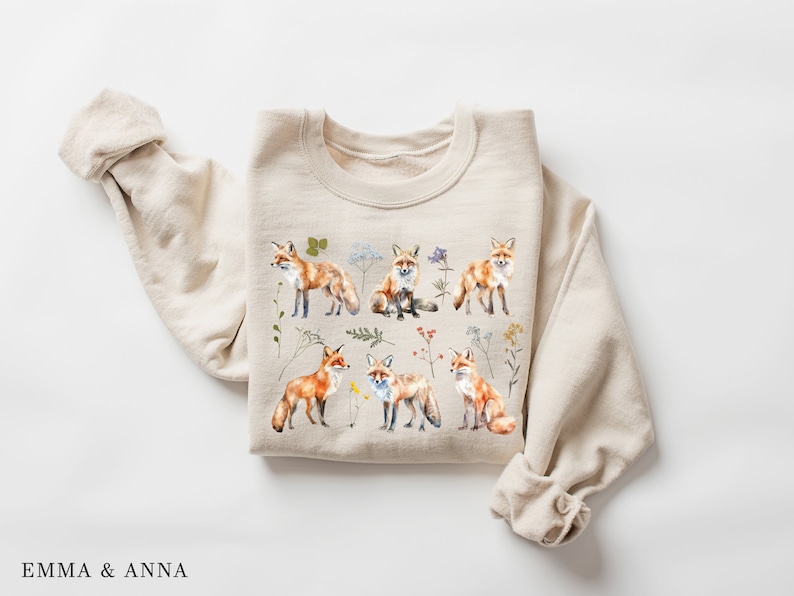May include: A beige sweatshirt with a print of multiple red foxes and wildflowers. The foxes are in various poses, including sitting, standing, and walking. The wildflowers are in shades of pink, purple, and yellow.
