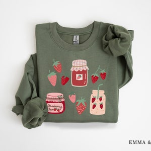 May include: A green sweatshirt with a strawberry jam and strawberry smoothie design. The design features jars of strawberry jam and a glass of strawberry smoothie with a straw, all surrounded by strawberries.